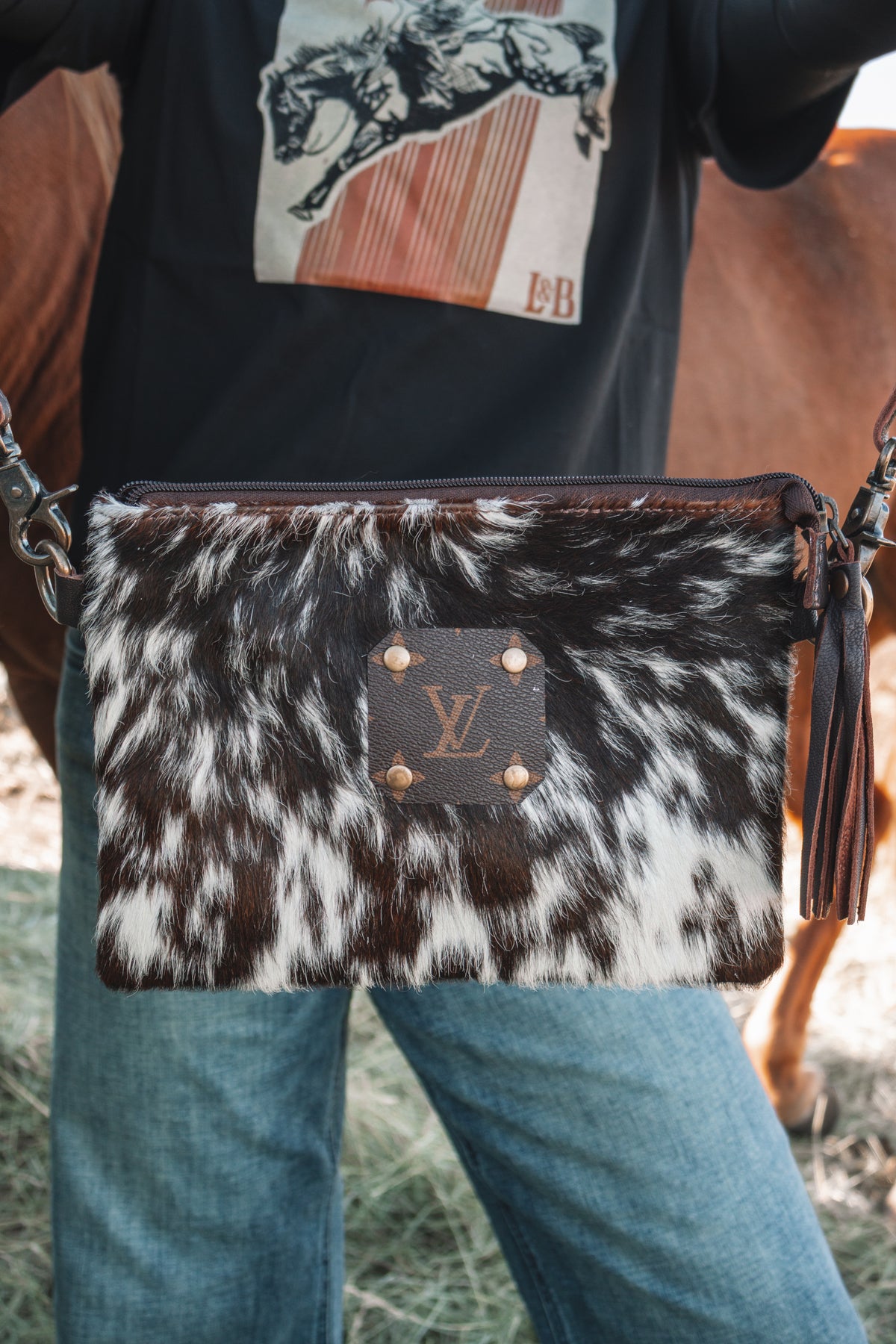 Brown and white cowhide crossbody purse with LV monogram accent and leather tassel, held in front of denim jeans and a black western graphic tee.