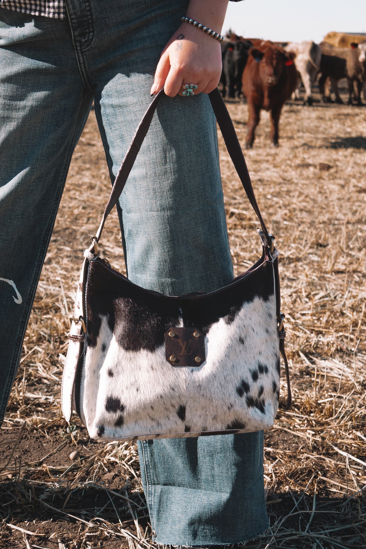 Black and white cowhide handbag with dark brown leather detailing and LV monogram accent, held by its shoulder strap in a pasture with cattle in the background.