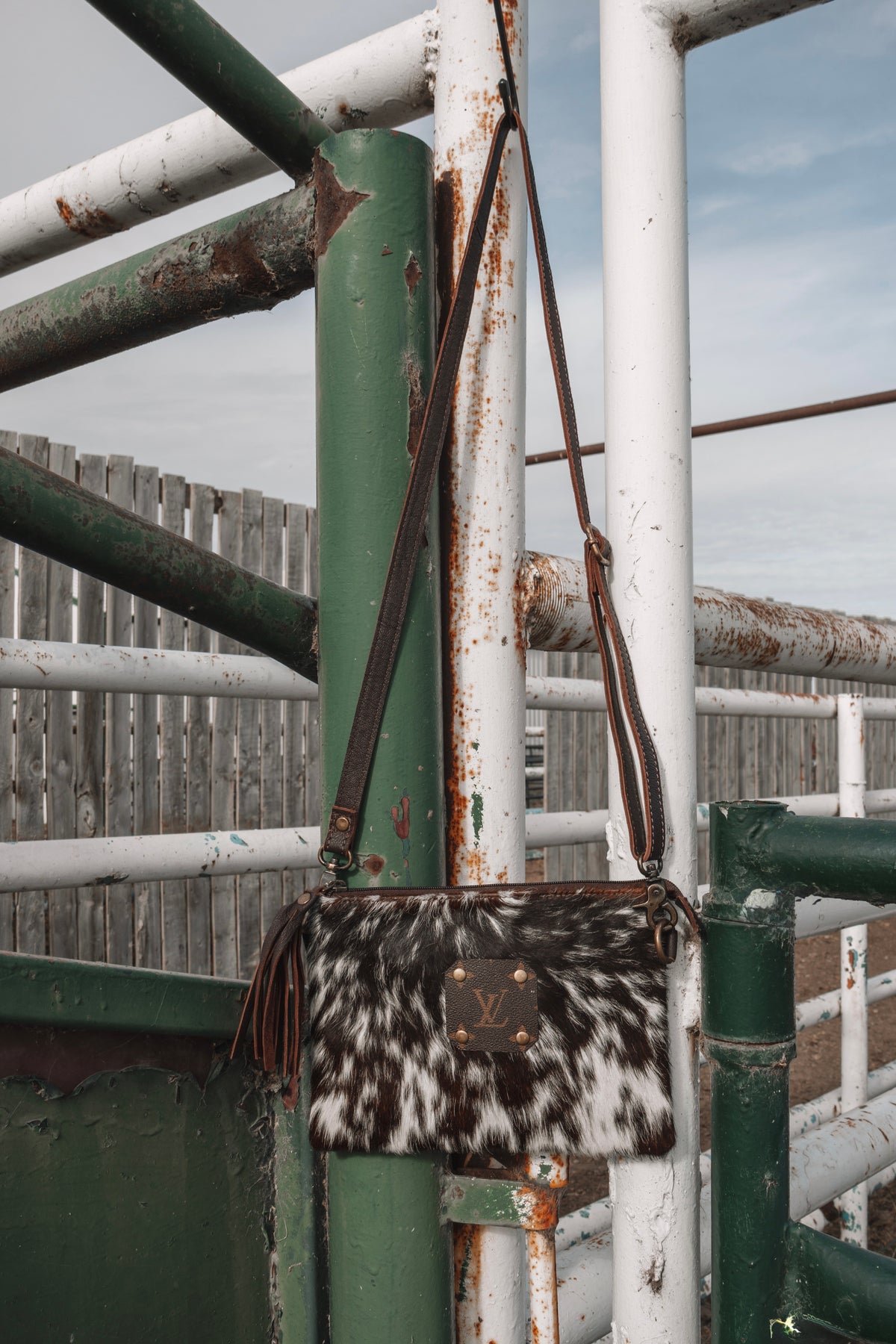 Brown and white cowhide crossbody bag with LV monogram patch, leather tassel, and long shoulder strap hanging on a rustic ranch gate.