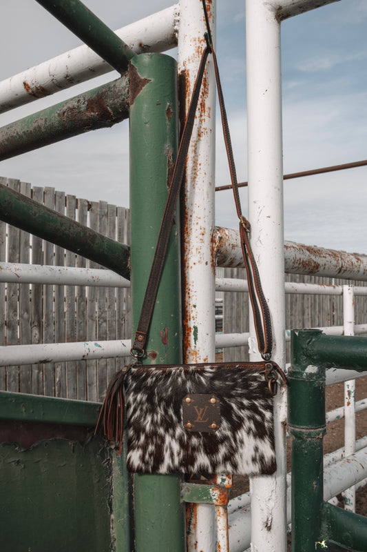 Brown and white cowhide crossbody bag with LV monogram patch, leather tassel, and long shoulder strap hanging on a rustic ranch gate.