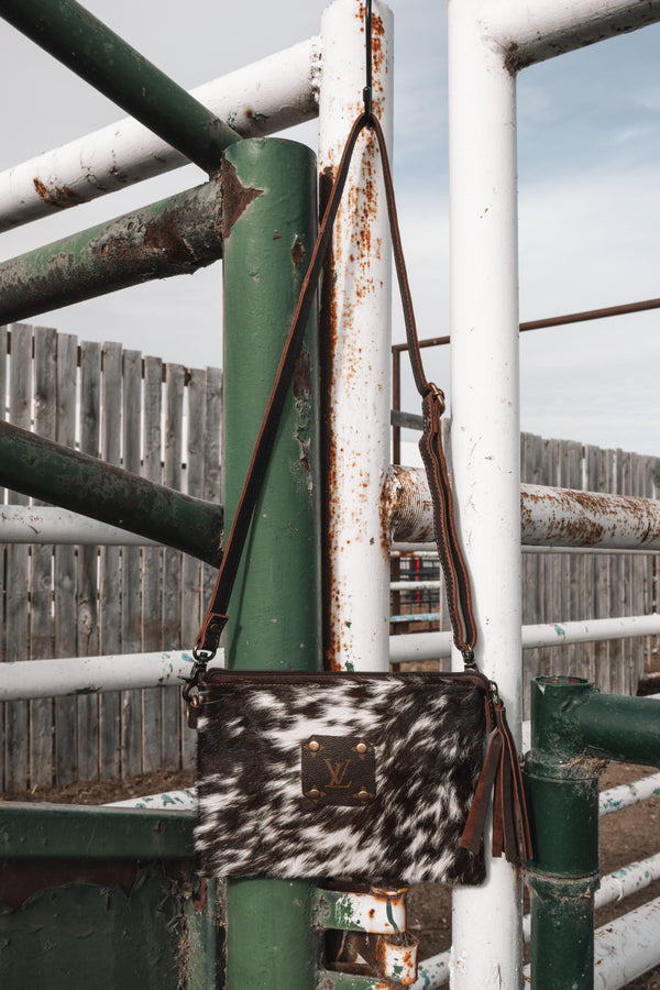 Brown and white speckled cowhide crossbody purse with LV monogram accent, adjustable leather strap, and tassel hanging on a green and white metal ranch gate.