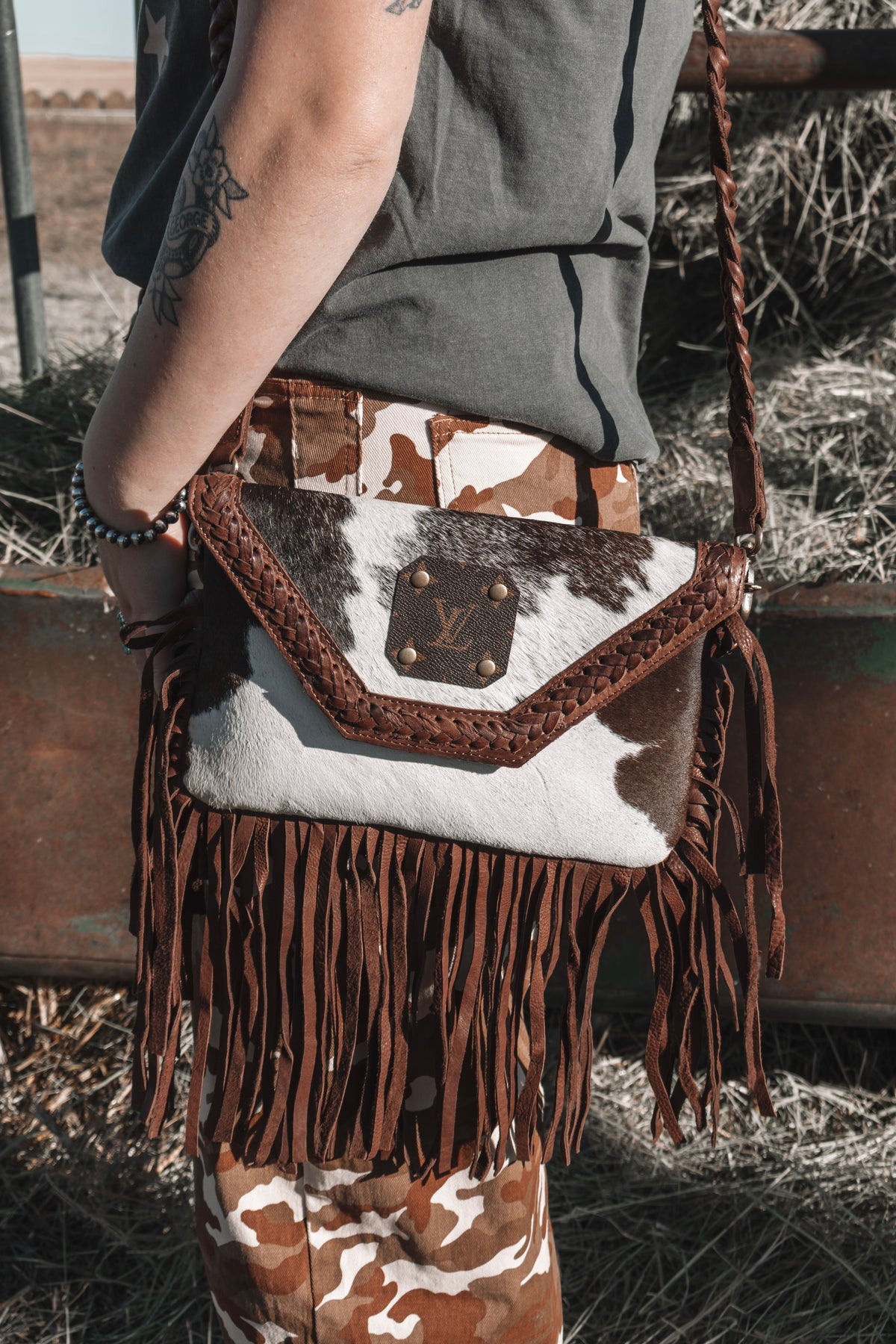 Model wearing a brown and white genuine cowhide crossbody bag with braided leather strap, long brown leather fringe and LV monogram patch, styled with brown camo pants.