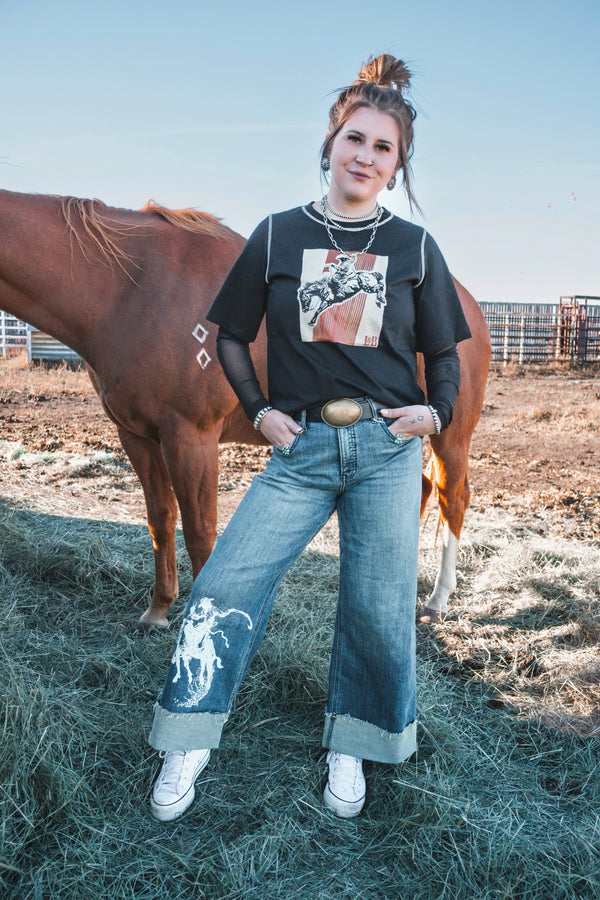 Woman wearing a black western bronc rider graphic tee layered over a black mesh long sleeve, paired with wide leg jeans, standing beside a horse.