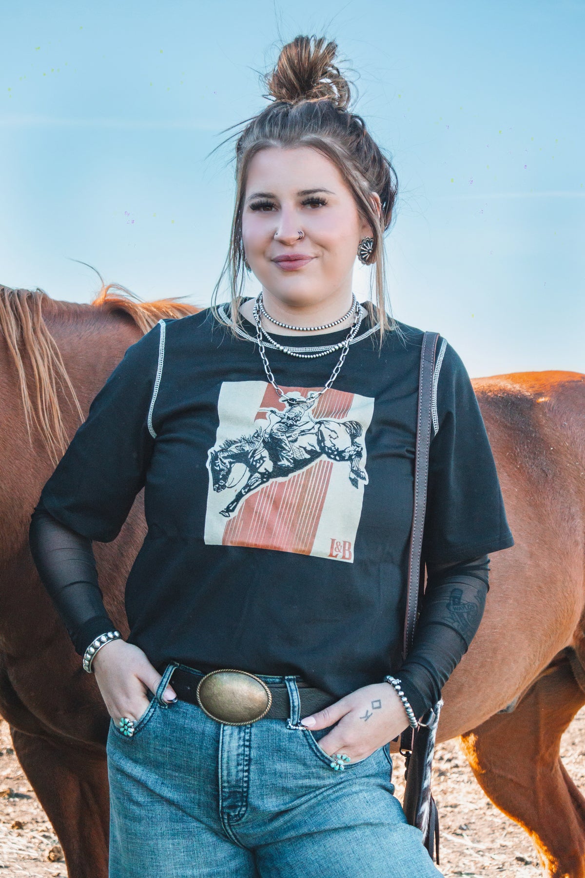 Woman wearing a black bronc rider graphic t-shirt with contrast stitching, layered over a black mesh long sleeve.