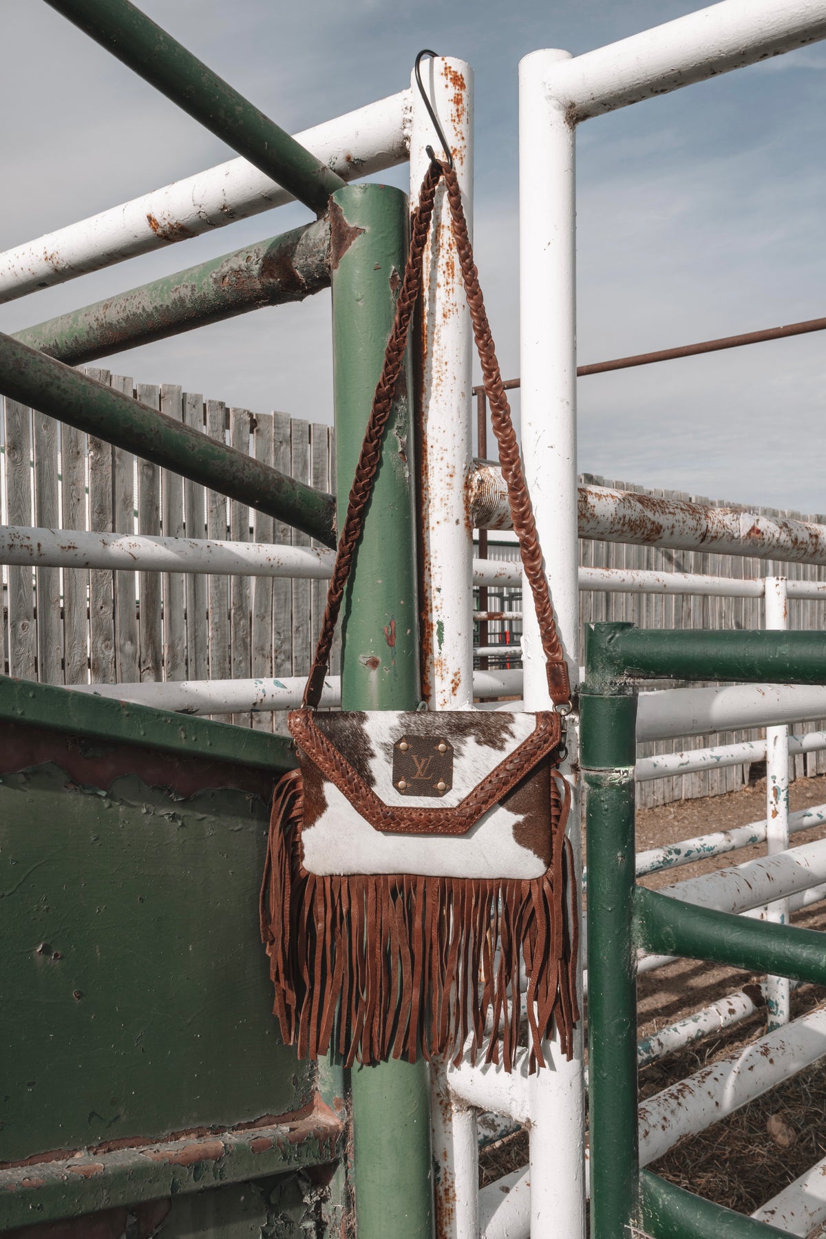 Brown and white genuine cowhide fringe bag with braided leather shoulder strap and long brown leather fringe, hanging on a ranch gate.