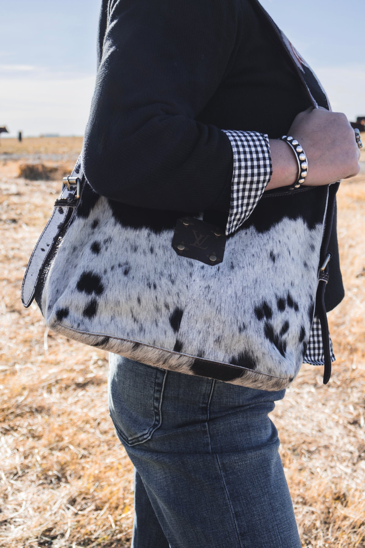 Black and white cowhide shoulder bag with dark brown leather strap and LV monogram patch, worn over the shoulder with a black sweater and denim jeans.