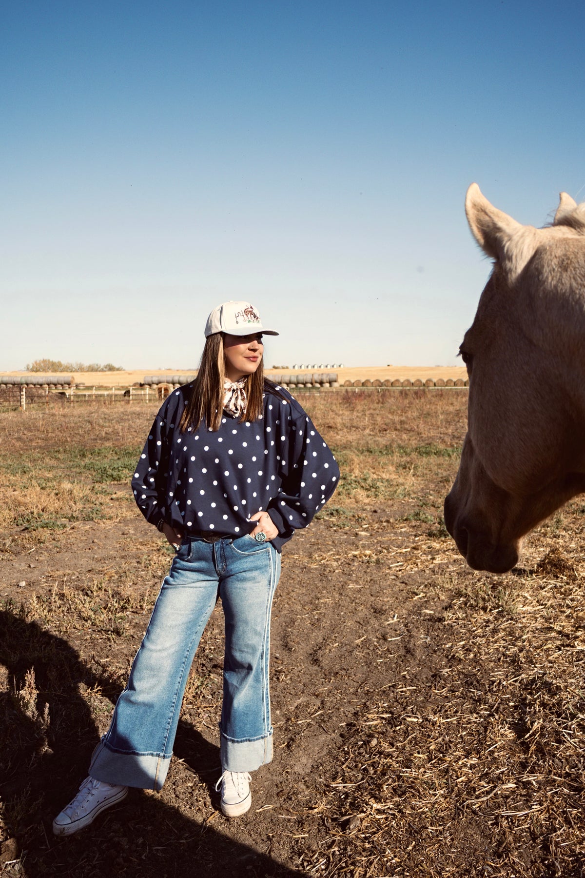 Women wearing a navy blue polka dot sweater, light wash wide-leg cuffed jeans and a white trucker hat besides a horse in pasture. Causal western boutique outfit from The Branded Roan