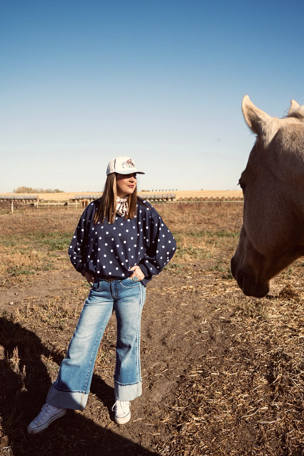 Women wearing a navy blue polka dot sweater, light wash wide-leg cuffed jeans and a white trucker hat besides a horse in pasture. Causal western boutique outfit from The Branded Roan