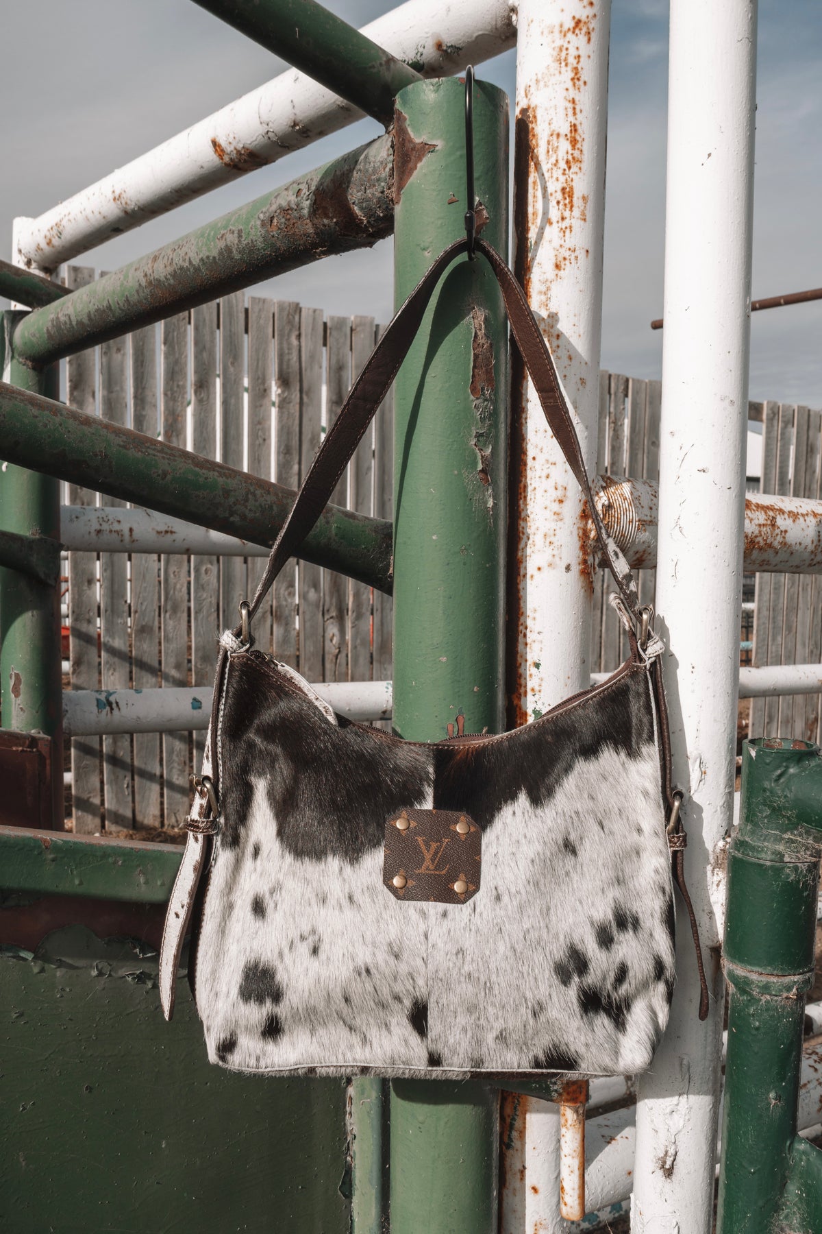 Black and white cowhide shoulder bag with dark brown leather strap and LV monogram accent, hanging on a rustic green and white ranch gate