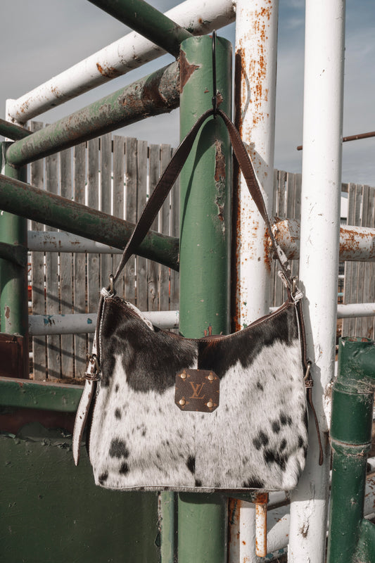 Black and white cowhide shoulder bag with dark brown leather strap and LV monogram accent, hanging on a rustic green and white ranch gate