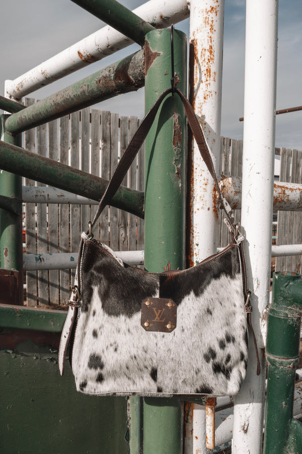 Black and white cowhide shoulder bag with dark brown leather strap and LV monogram accent, hanging on a rustic green and white ranch gate