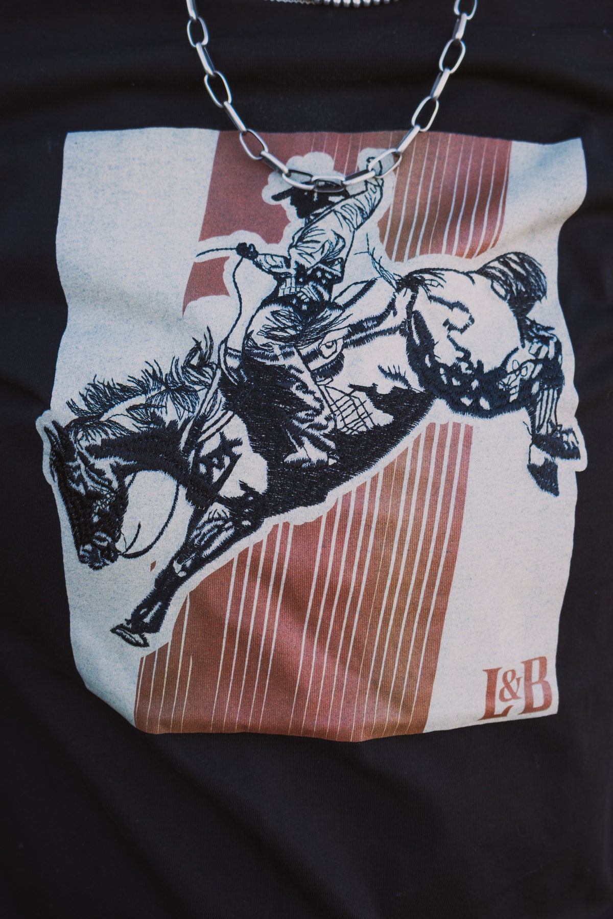 Close-up of black western graphic tee featuring a detailed bronc rider illustration with rust-coloured stripes and a cream background.
