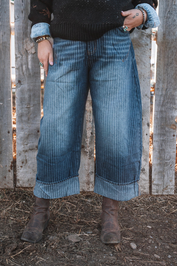 Women's vintage wash blue pinstripe wide leg jeans featuring oversized rolled cuffs, styled with a black knit sweater and brown western ankle boots.