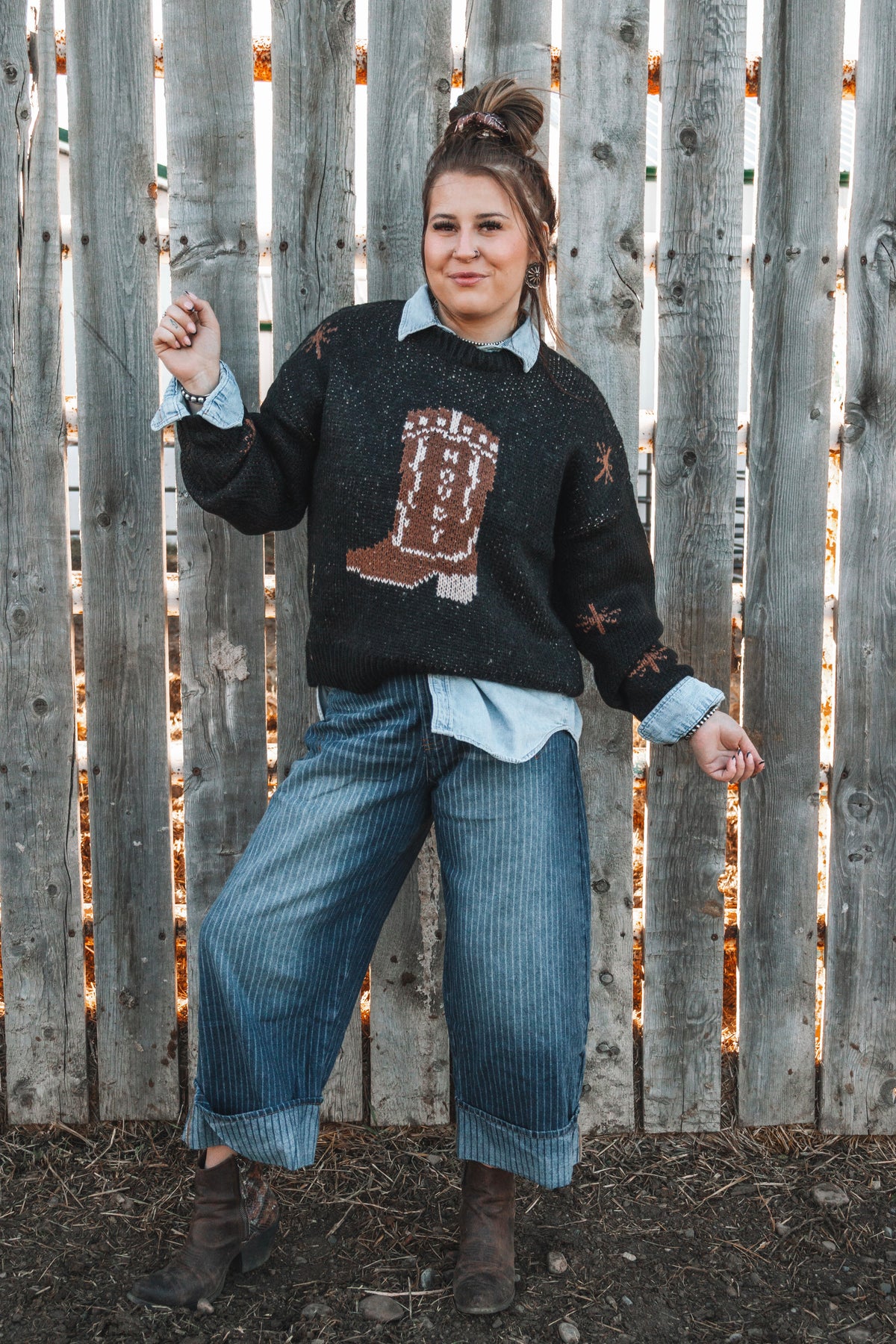 Women's black western knit sweater featuring a brown cowboy boot graphic, styled over a light wash denim shirt with striped wide leg jeans and brown ankle boots.