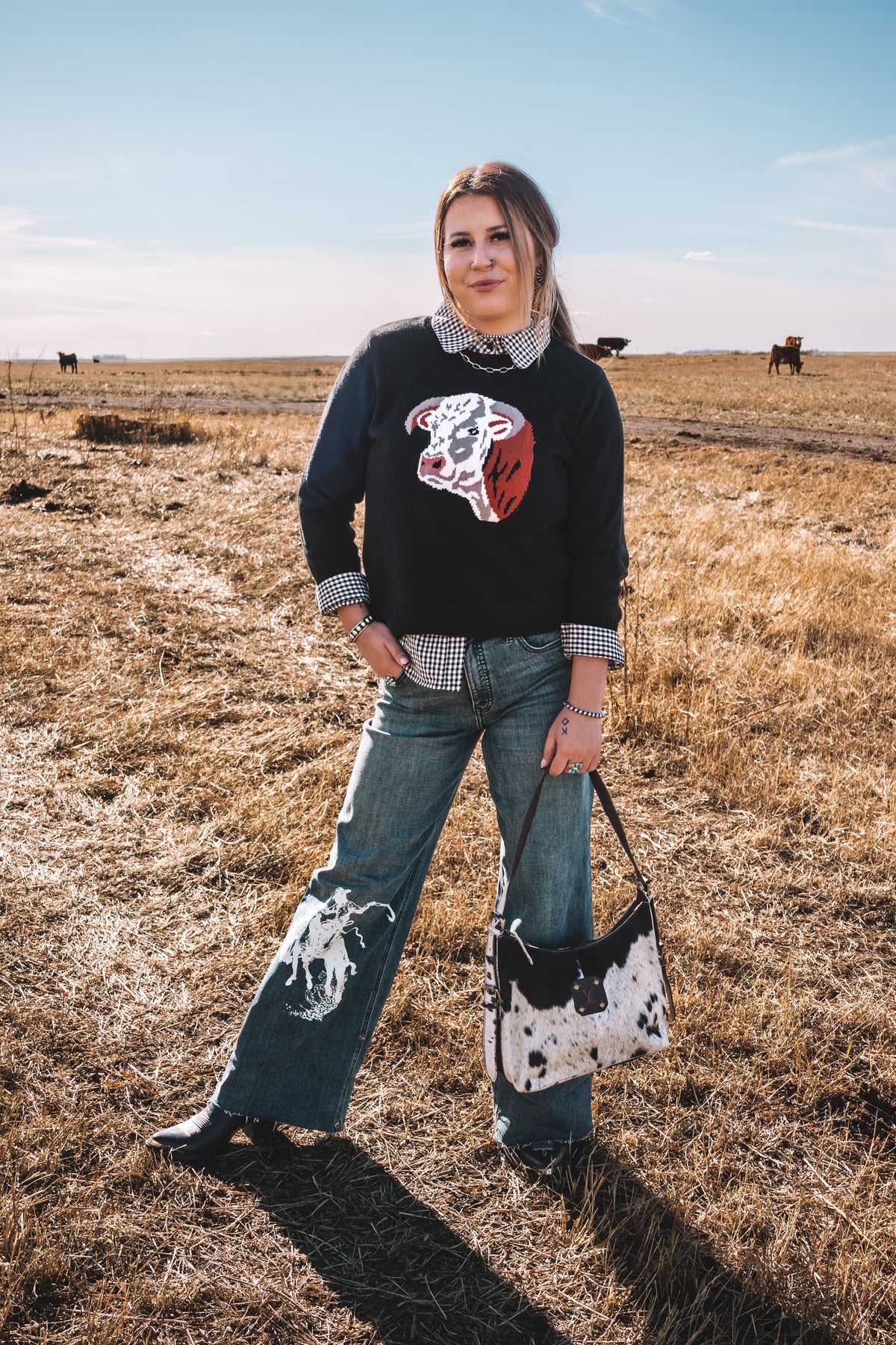 Women's black western knit sweater featuring a Hereford cow graphic on the front, styled with a black and white gingham collared shirt and wide leg denim jeans.