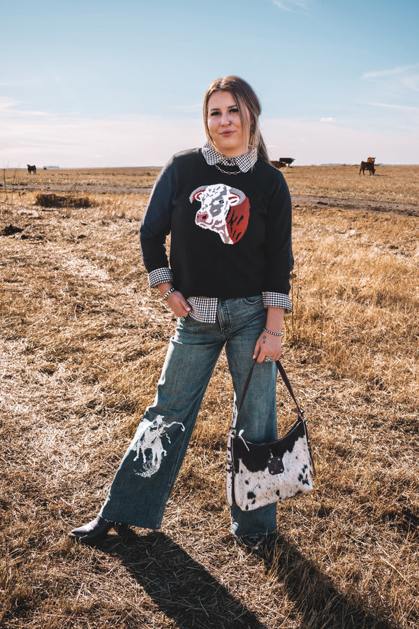 Women's black western knit sweater featuring a Hereford cow graphic on the front, styled with a black and white gingham collared shirt and wide leg denim jeans.