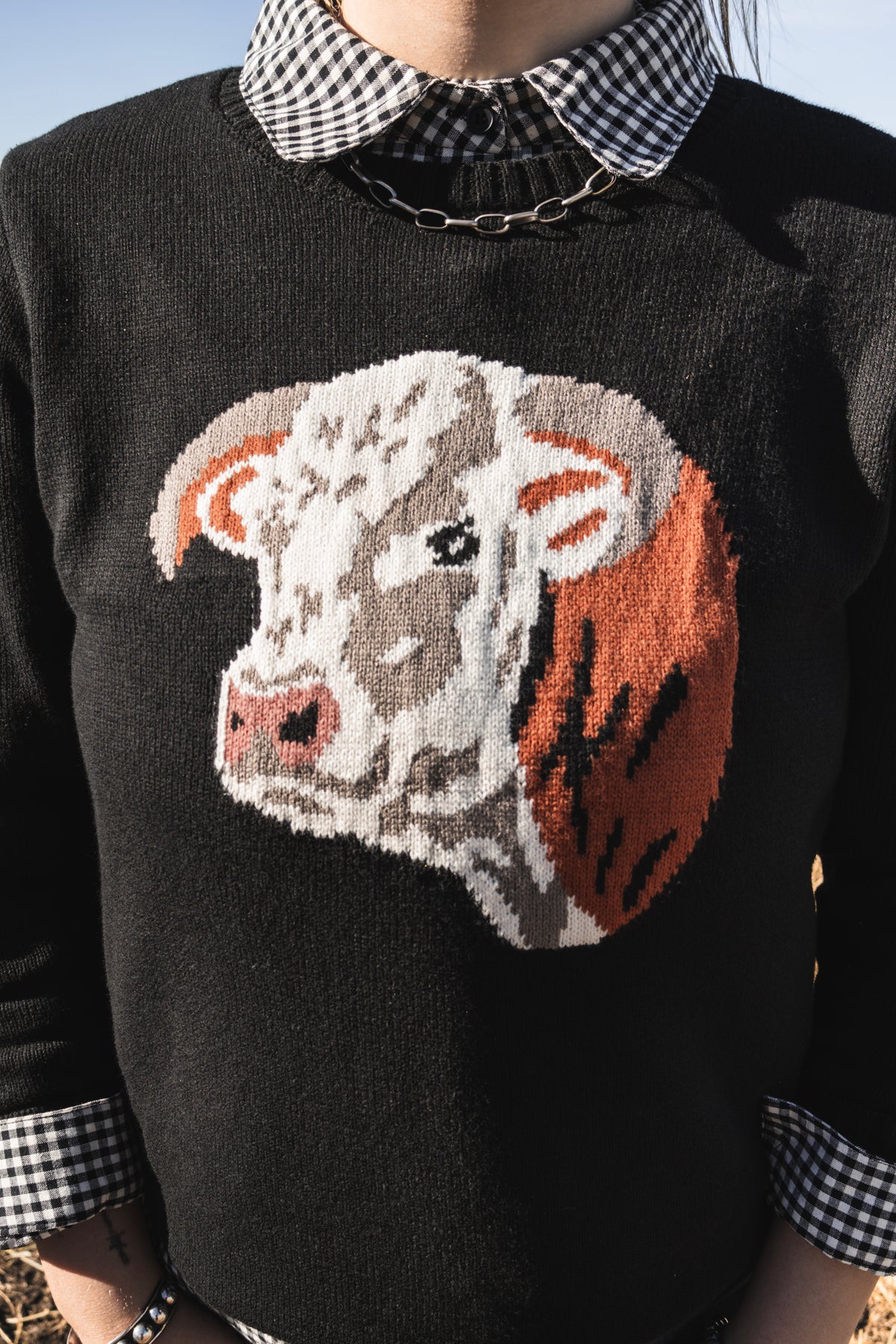 Women's black western knit sweater featuring a Hereford cow graphic on the front, styled with a black and white gingham collared shirt and wide leg denim jeans.