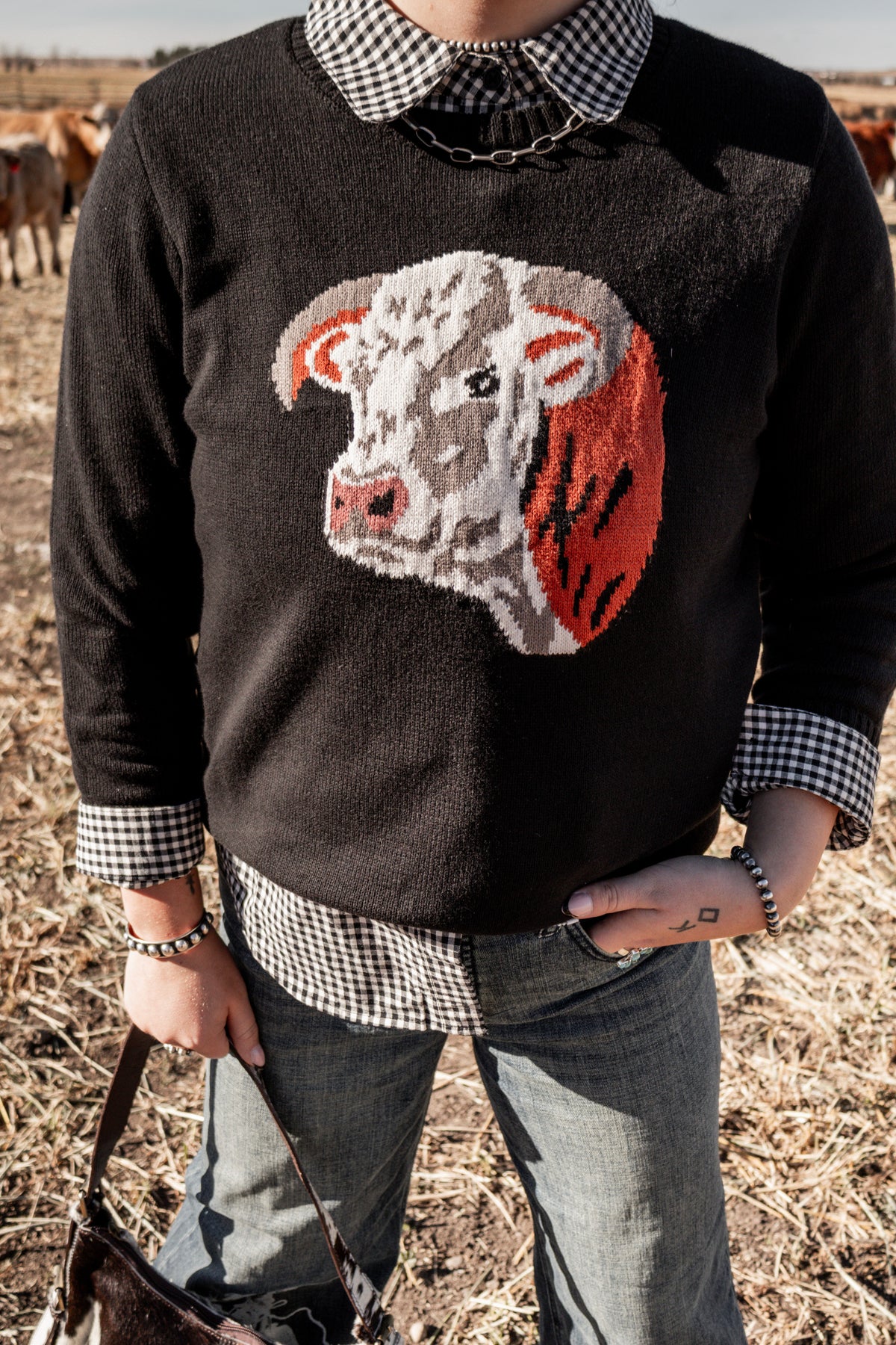 Women's black western knit sweater featuring a Hereford cow graphic on the front, styled with a black and white gingham collared shirt and wide leg denim jeans.