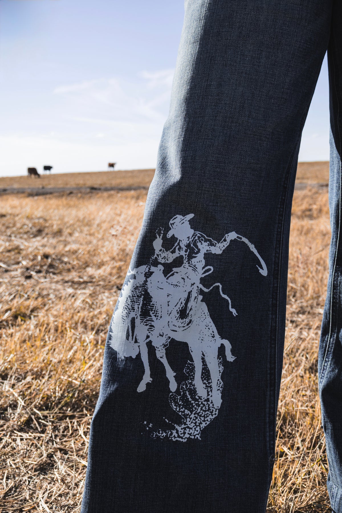 Close-up of women's western wide leg jeans featuring a white bronc rider graphic on medium wash stretch denim, styled uncuffed with cowboy boots.