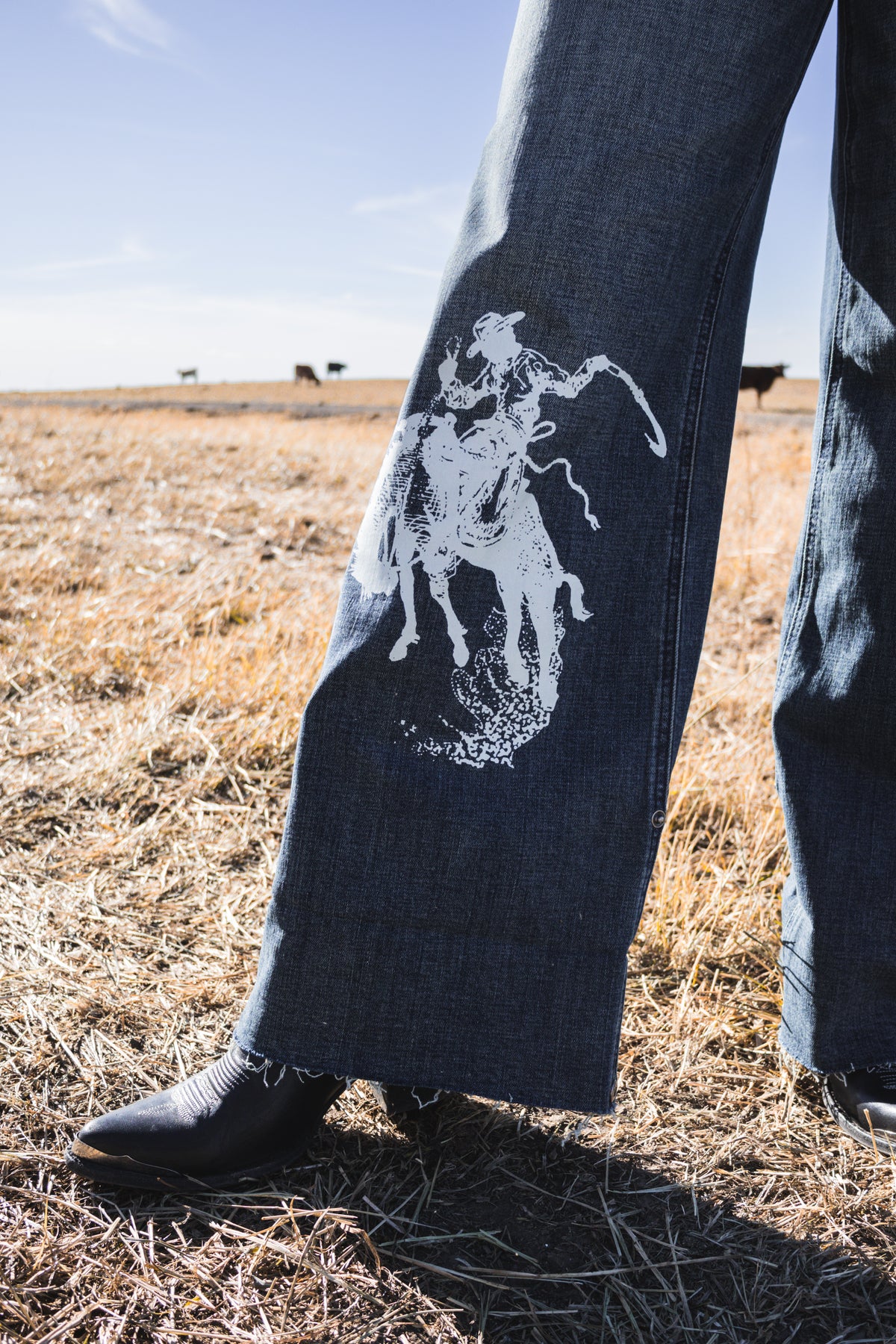 Close-up of women's western wide leg jeans featuring a white bronc rider graphic on medium wash stretch denim, styled uncuffed with cowboy boots.