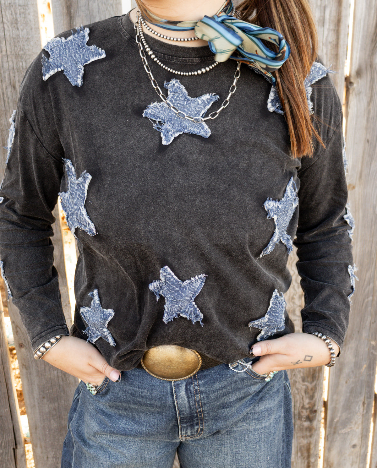 Close-up of women's vintage black long sleeve top featuring distressed blue denim star patches with frayed edges, styled with western jewelry and blue jeans.