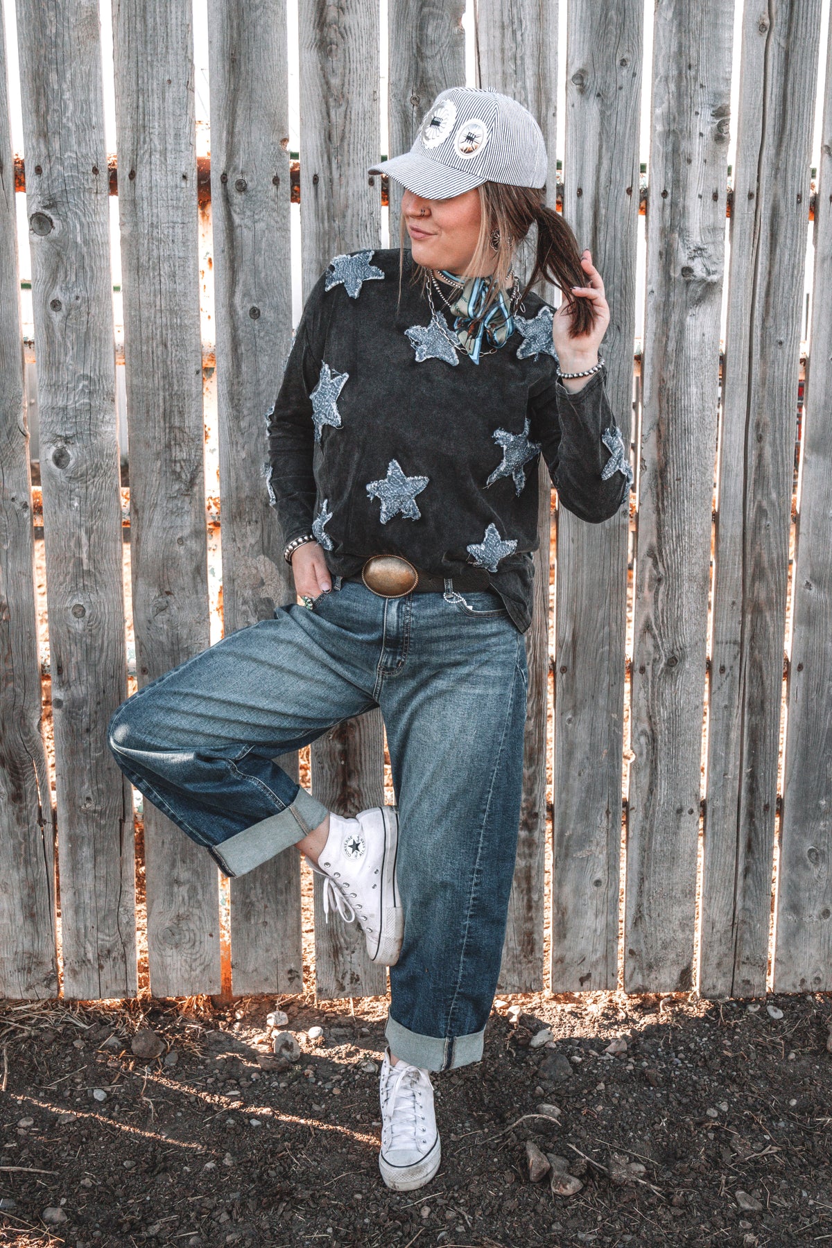 Women's vintage washed black long sleeve top featuring blue denim star patches, styled with cuffed wide leg jeans, white sneakers, a western belt and a baseball cap.