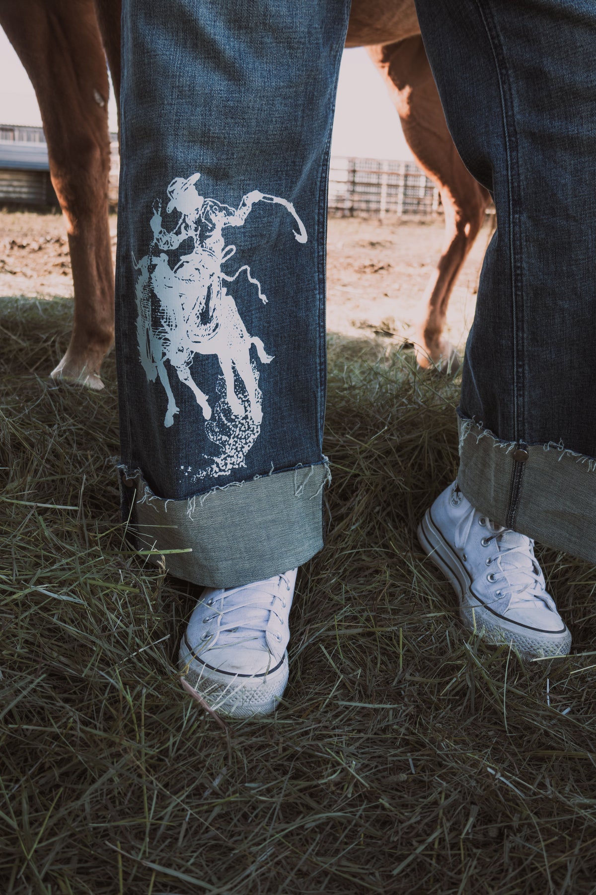 Close-up of women's high rise western wide leg jeans in medium stonewash denim, featuring a white bronc rider graphic and adjustable button cuffs, styled with white sneakers.