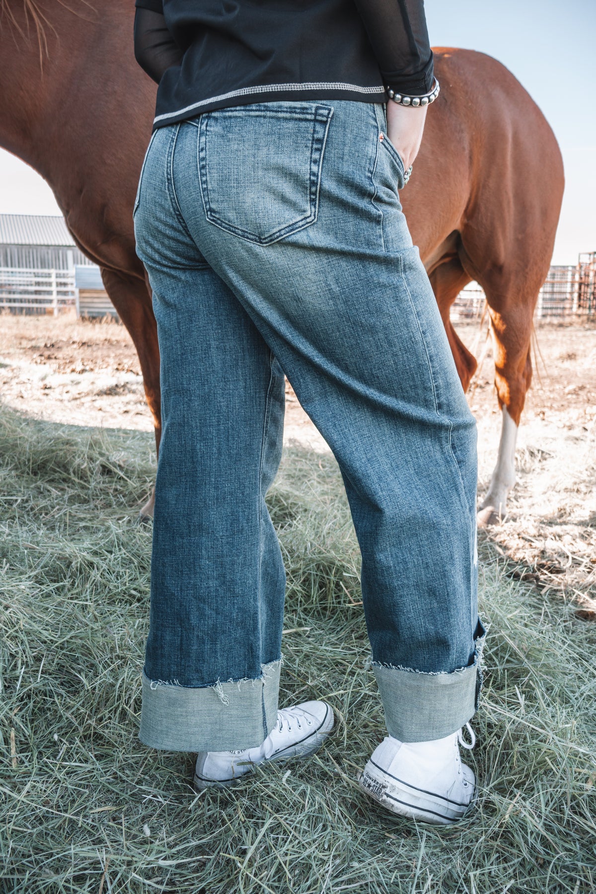 Back view of women's high rise western wide leg jeans in medium stonewash denim, featuring classic back pockets, a relaxed fit, and adjustable rolled button cuffs, styled with white sneakers.
