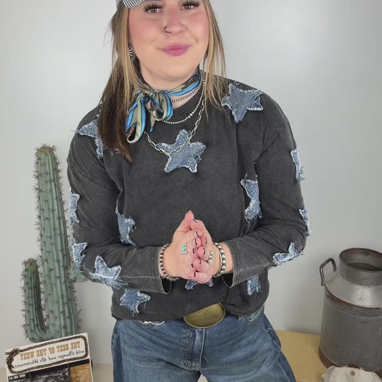Women's vintage washed black long sleeve top featuring distressed blue denim star patches throughout, styled with wide leg jeans, a western belt, turquoise neck scarf and pinstripe baseball cap.