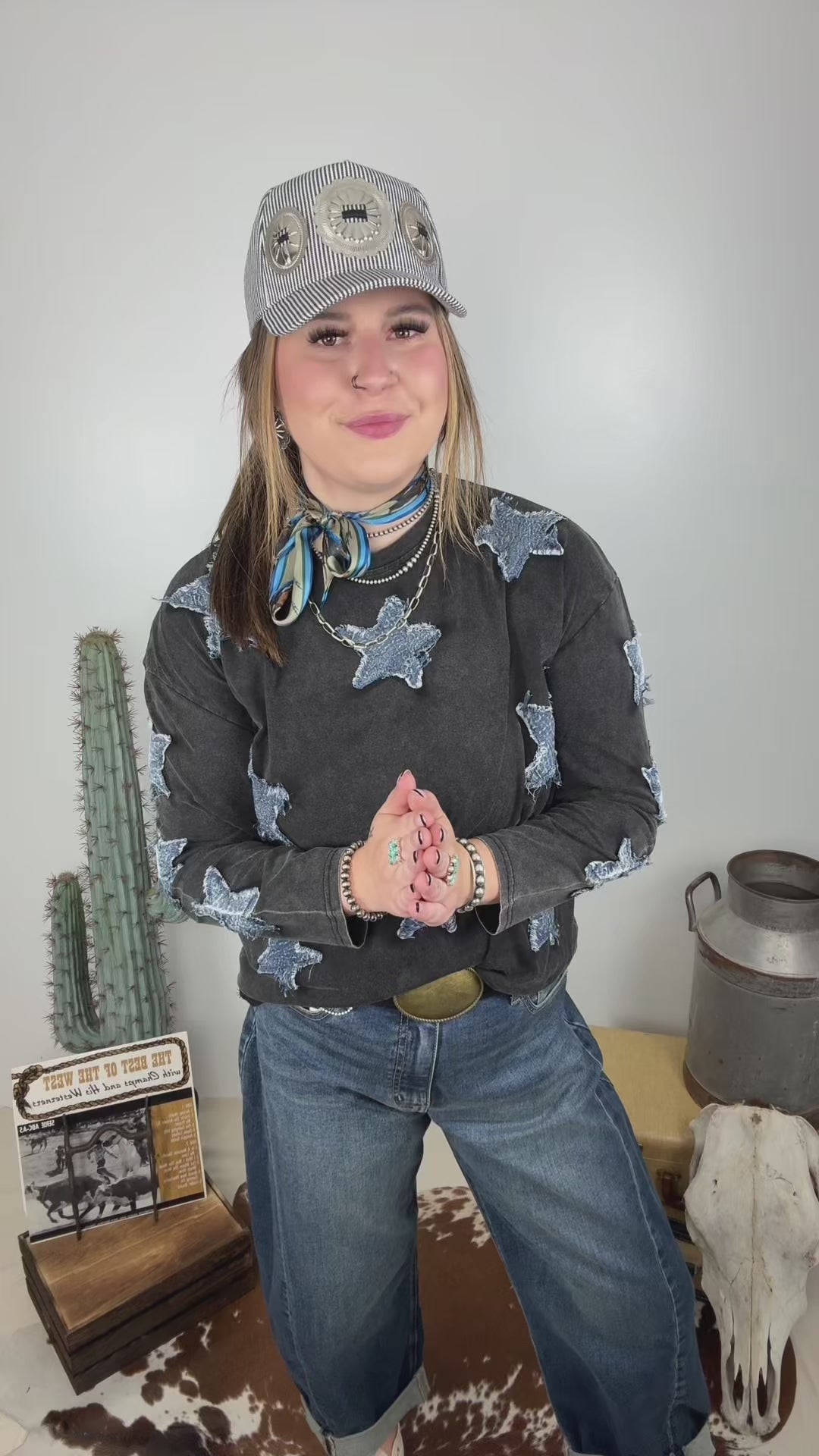 Women's vintage washed black long sleeve top featuring distressed blue denim star patches throughout, styled with wide leg jeans, a western belt, turquoise neck scarf and pinstripe baseball cap.