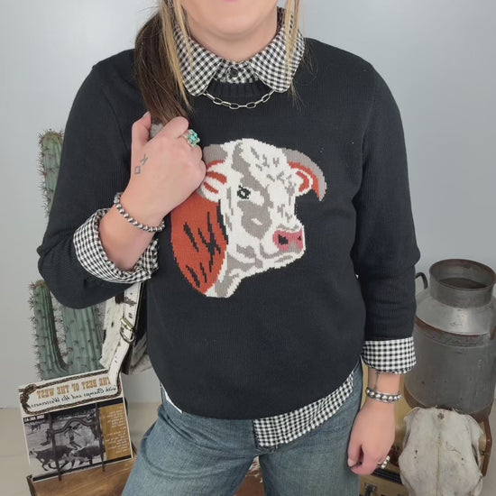 Women's black western knit sweater featuring a Hereford cow graphic on the front, styled with a black and white gingham collared shirt and wide leg denim jeans.