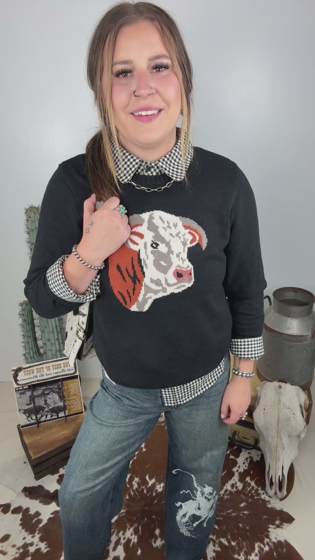 Women's black western knit sweater featuring a Hereford cow graphic on the front, styled with a black and white gingham collared shirt and wide leg denim jeans.